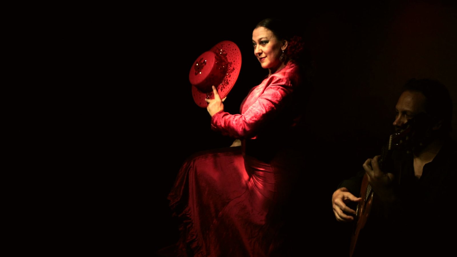 La Choni, vêtue de rouge et son chapeau à la main, avec le guitariste à ses côtés sur le fond noir de la scène de CasaLa Teatro