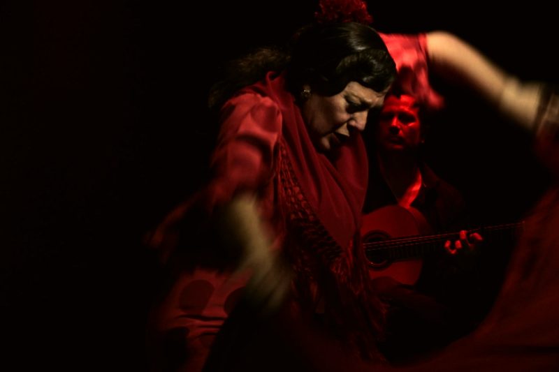 La Choni, de rojo y con una flor en el pelo, en pleno baile con el mantón; detrás, el guitarrista bajo la luz roja