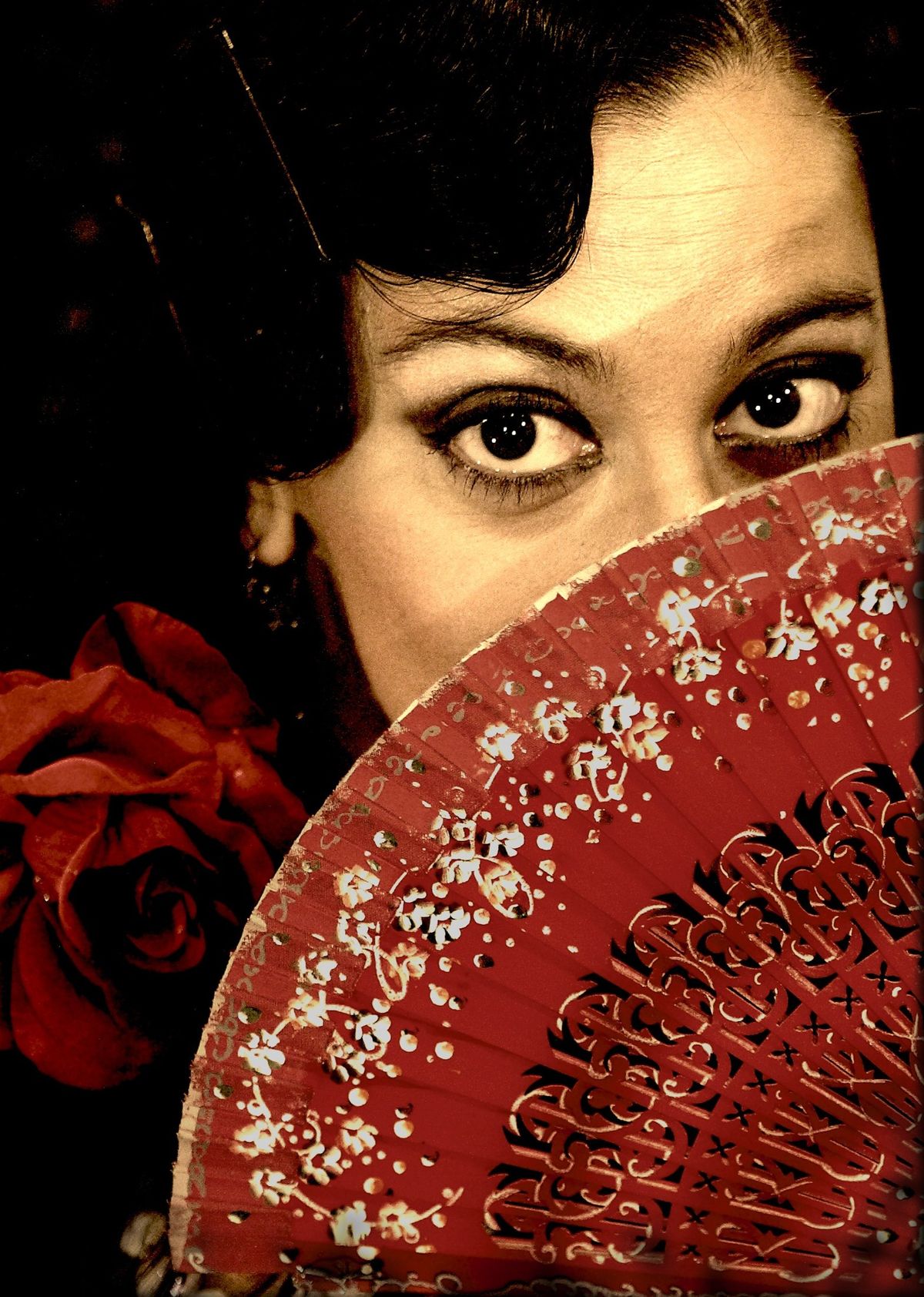 Rostro de una bailaora asomando por encima de un abanico rojo de flamenco, con una rosa roja junto al pelo