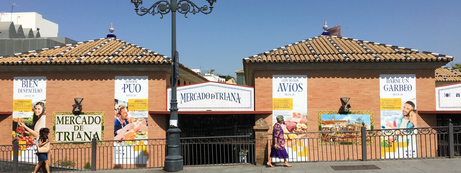 La fachada del Mercado de Triana desde la calle, con el rótulo de azulejos sobre la entrada, los tejados de teja y dos mujeres pasando por la acera