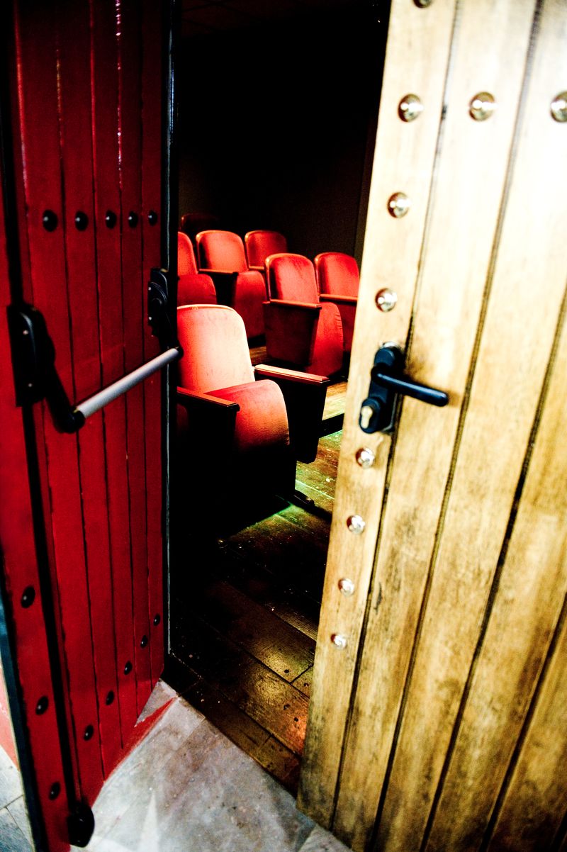 Las butacas rojas de la sala vistas por el hueco de la puerta entreabierta del teatro, entre una hoja de madera roja y otra clara